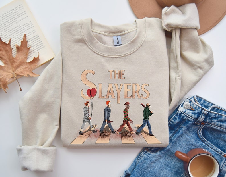 May include: A beige sweatshirt with a graphic print of horror movie characters walking across a crosswalk. The text "The Slayers" is printed above the characters.
