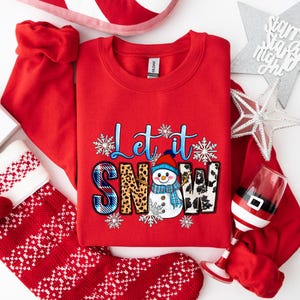 Op de afbeelding: Rood sweatshirt met een sneeuwpop-afbeelding en de tekst "Let it Snow". De sneeuwpop draagt ​​een blauwe muts en sjaal en houdt een stok vast. De tekst is in wit, blauw en zwart lettertype.