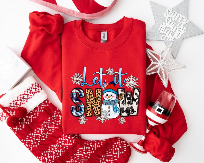 Op de afbeelding: Rood sweatshirt met een sneeuwpop-afbeelding en de tekst "Let it Snow". De sneeuwpop draagt ​​een blauwe muts en sjaal en houdt een stok vast. De tekst is in wit, blauw en zwart lettertype.