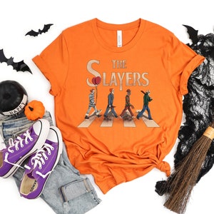 May include: Orange t-shirt with a graphic of horror movie characters walking across a crosswalk. The text "The Slayers" is above the characters.
