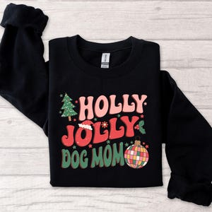 May include: Black sweatshirt with a Christmas-themed design. The text reads "Holly Jolly Dog Mom" in a retro font. The design also includes a Christmas tree, a Santa hat, and a disco ball ornament.