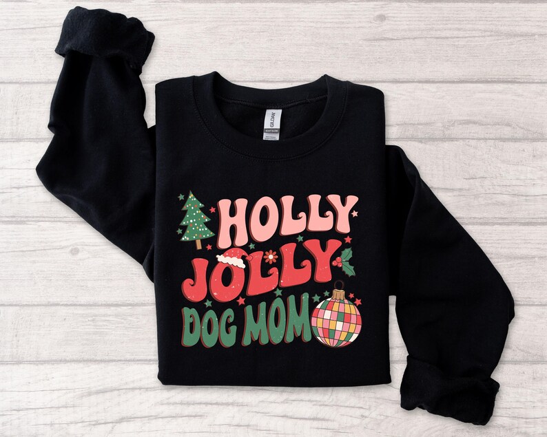 May include: Black sweatshirt with a Christmas-themed design. The text reads "Holly Jolly Dog Mom" in a retro font. The design also includes a Christmas tree, a Santa hat, and a disco ball ornament.