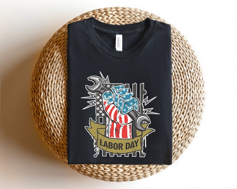 May include: Black t-shirt with a graphic of a hand holding a wrench. The hand is painted in the colors of the American flag and the wrench is silver. The graphic is surrounded by stars and the text "Labor Day".