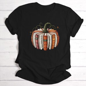 May include: A black short-sleeve t-shirt featuring a central graphic of a pumpkin constructed from the spines and pages of various books. The pumpkin has a green stem with curling vines and displays books in shades of orange, red, and brown.