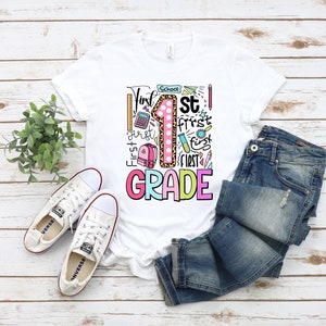 May include: White t-shirt with a colorful "First Grade" graphic. The design includes a large number 1 with a leopard print pattern, school supplies, and the words "First" and "Grade". The shirt is paired with blue jeans and white sneakers.