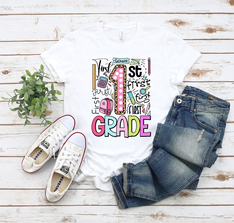 May include: White t-shirt with a colorful "First Grade" graphic. The design includes a large number 1 with a leopard print pattern, school supplies, and the words "First" and "Grade". The shirt is paired with blue jeans and white sneakers.