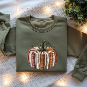 May include: An olive green crewneck sweatshirt featuring a central graphic of a pumpkin constructed from stacked, open books with colorful spines and a green stem. The sweatshirt has long sleeves with ribbed cuffs and a ribbed neckline.