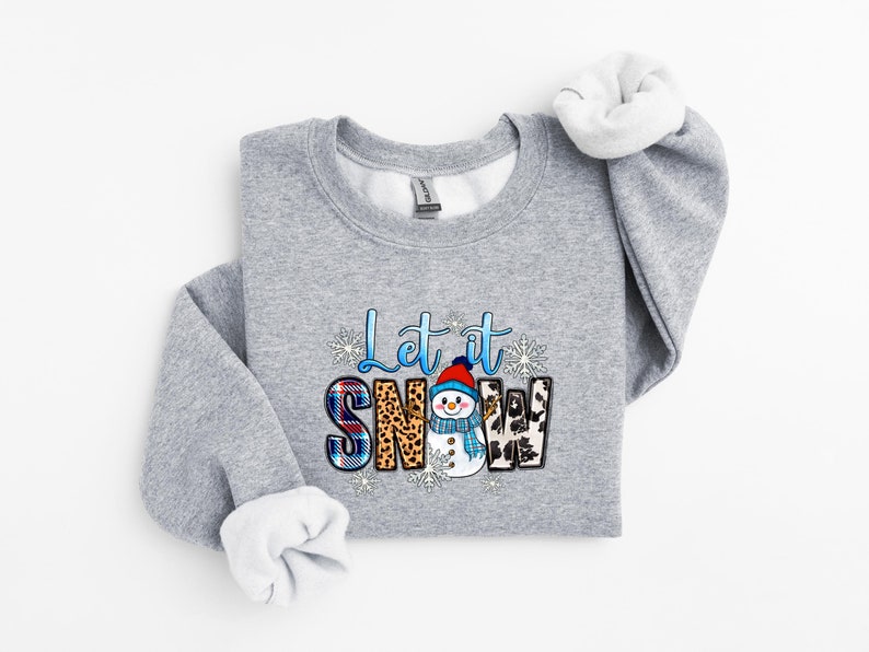 Op de afbeelding: Een grijs sweatshirt met een feestelijk ontwerp met een sneeuwpop en de tekst "Let it Snow". De letters zijn versierd met ruitjes, luipaardprint en andere patronen.