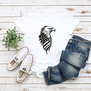May include: White t-shirt with a black and white graphic of an eagle with the American flag on its chest.