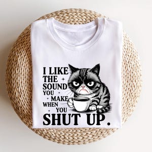 May include: White t-shirt featuring a grumpy cat graphic holding a coffee cup. The cat is black and gray with an angry expression. Text on the shirt reads, "I LIKE THE SOUND YOU MAKE WHEN YOU SHUT UP."