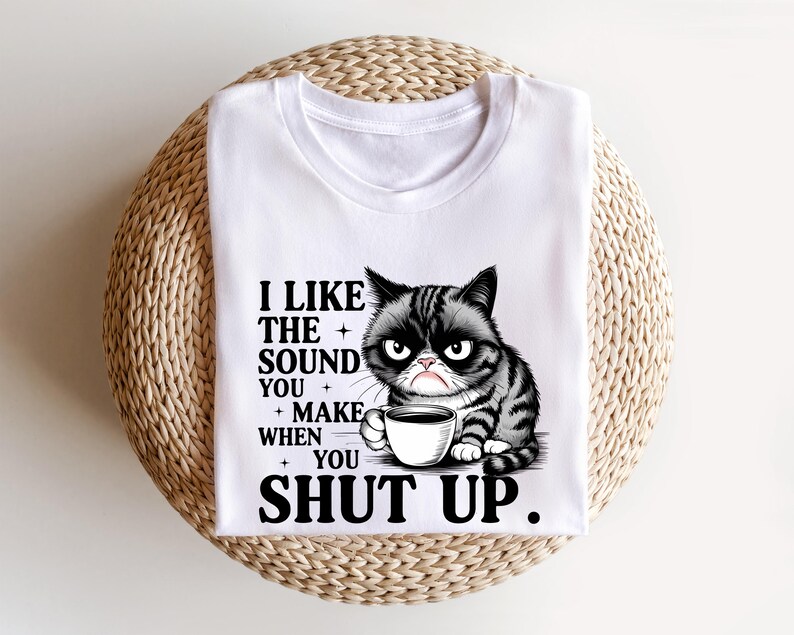 May include: White t-shirt featuring a grumpy cat graphic holding a coffee cup. The cat is black and gray with an angry expression. Text on the shirt reads, "I LIKE THE SOUND YOU MAKE WHEN YOU SHUT UP."