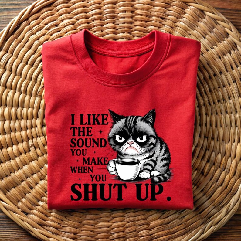 May include: A red t-shirt featuring a grumpy cat illustration holding a coffee cup. The shirt has the text "I LIKE THE SOUND YOU MAKE WHEN YOU SHUT UP" in black. The cat is black and gray with an angry expression.