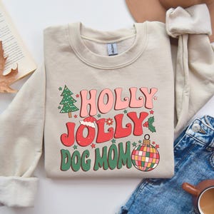 May include: A beige sweatshirt with a Christmas-themed design. The text reads "Holly Jolly Dog Mom" with a red and green ornament, a Christmas tree, and a Santa hat.