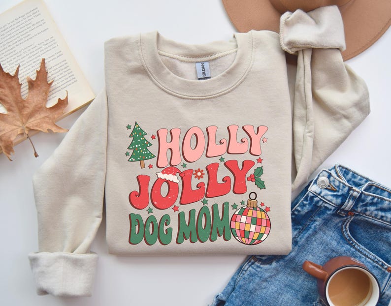 May include: A beige sweatshirt with a Christmas-themed design. The text reads "Holly Jolly Dog Mom" with a red and green ornament, a Christmas tree, and a Santa hat.