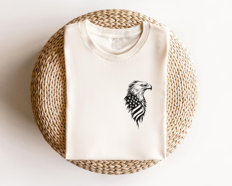 May include: A white t-shirt with a black and white image of an eagle with an American flag design on its chest.