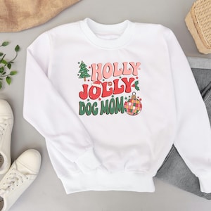 May include: White sweatshirt with a Christmas-themed graphic that says "Holly Jolly Dog Mom" in pink, red, and green lettering. The graphic also includes a Christmas tree, a Santa hat, and a disco ball ornament.