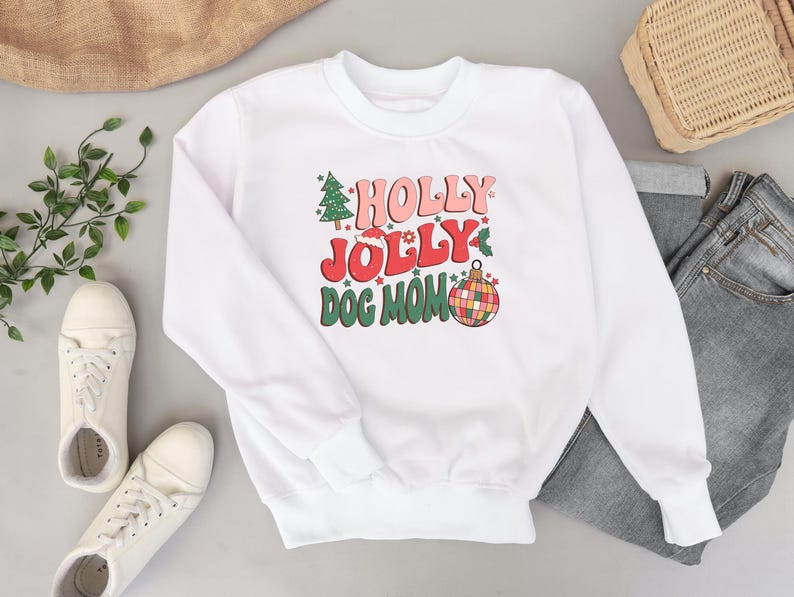 May include: White sweatshirt with a Christmas-themed graphic that says "Holly Jolly Dog Mom" in pink, red, and green lettering. The graphic also includes a Christmas tree, a Santa hat, and a disco ball ornament.