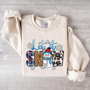 Op de afbeelding: Een cr&egrave;mekleurige trui met een grafisch ontwerp van een sneeuwpop en de tekst "Let it Snow". De sneeuwpop draagt een blauw-witte sjaal en een rode muts. De tekst is in blauw en wit.