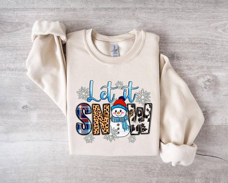 Op de afbeelding: Een cr&egrave;mekleurige trui met een grafisch ontwerp van een sneeuwpop en de tekst "Let it Snow". De sneeuwpop draagt een blauw-witte sjaal en een rode muts. De tekst is in blauw en wit.