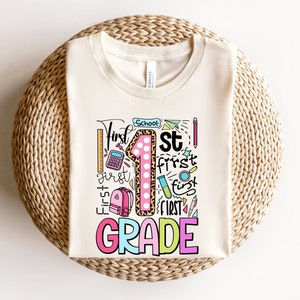 May include: Cream-colored t-shirt with a colorful graphic design for first grade. The design includes the words "First Grade" in a playful font, a large number "1" with a leopard print pattern, and school-themed illustrations.