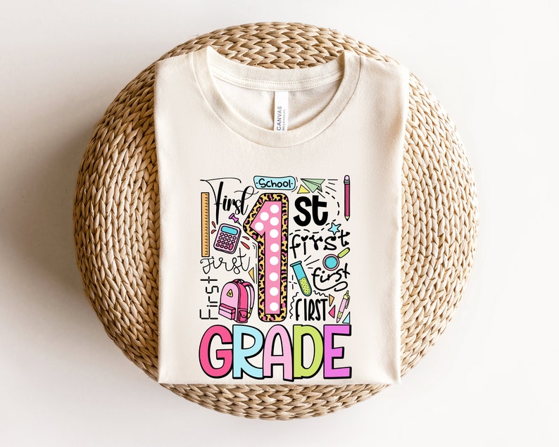 May include: Cream-colored t-shirt with a colorful graphic design for first grade. The design includes the words "First Grade" in a playful font, a large number "1" with a leopard print pattern, and school-themed illustrations.