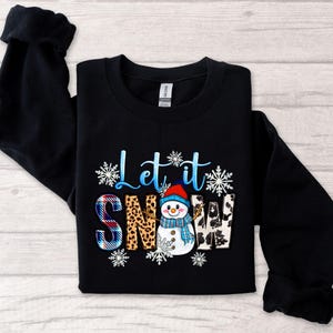 Op de afbeelding: Zwart sweatshirt met een sneeuwpop-afbeelding en de tekst "Let it snow". De sneeuwpop draagt een blauwe en witte muts en sjaal. De tekst is in wit en blauw.