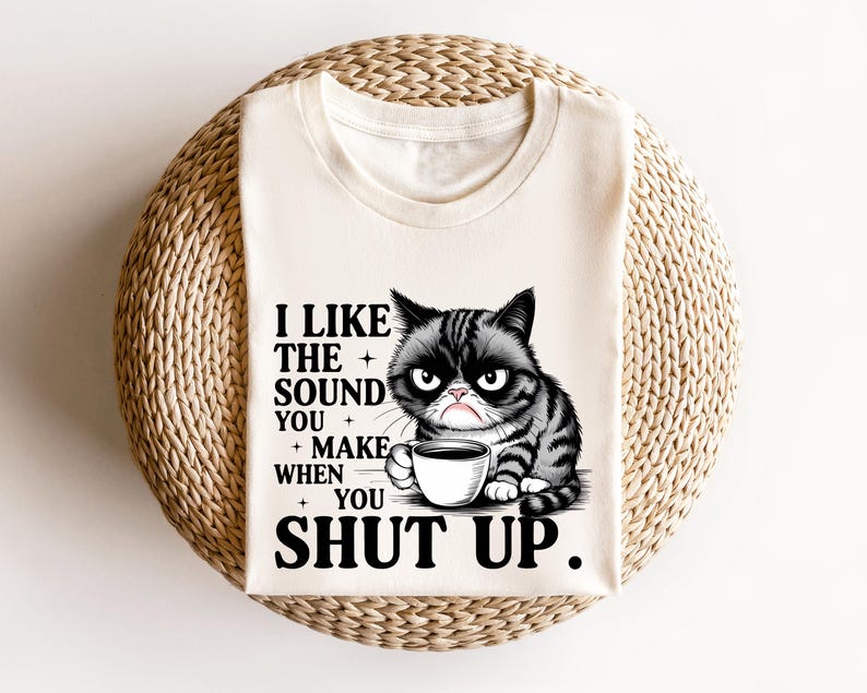 May include: Cream-colored t-shirt featuring a grumpy cat illustration with a coffee cup. The text reads, "I LIKE THE SOUND YOU MAKE WHEN YOU SHUT UP." The cat is black and gray with a displeased expression.