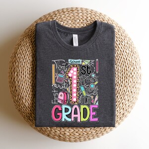 May include: Dark gray t-shirt with a colorful graphic design for first grade. The design includes a large number 1 with a leopard print pattern, school supplies, and the words "First Grade".