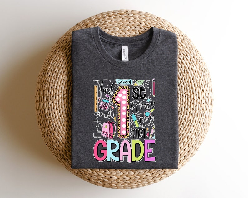 May include: Dark gray t-shirt with a colorful graphic design for first grade. The design includes a large number 1 with a leopard print pattern, school supplies, and the words "First Grade".