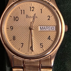 May include: Gold-toned Bulova wristwatch with a textured dial, featuring the date "MAR 10". The watch has a gold band and a round face with gold hands and hour markers. The watch face displays the Bulova logo.
