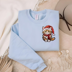 Op de afbeelding: Een lichtblauw sweatshirt met een geborduurde kat in rood en oranje die een kerstmanmuts en een rode sjaal draagt.