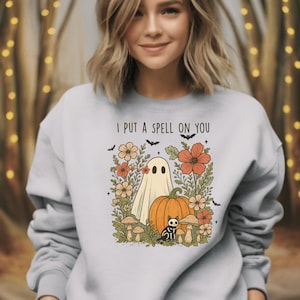 Op de afbeelding: Een lichtgrijs sweatshirt met een Halloween-thema. De afbeelding toont een spook, pompoen, zwarte kat, bloemen en de tekst "I PUT A SPELL ON YOU". Het ontwerp gebruikt oranje, beige en zwarte kleuren.