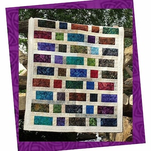 May include: A colorful quilt with a brick-like pattern, featuring rectangles in shades of red, blue, green, and purple. The quilt is framed in a purple border with the word "Jericho" at the top. The quilt is hanging on a wooden structure.