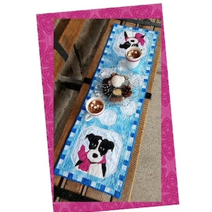 Pode incluir: Um corredor de mesa acolchoado azul e branco com um design de cão preto e branco e o texto "Hope in January". O corredor está sobre uma mesa de madeira com uma caneca de chocolate quente e um prato de biscoitos.