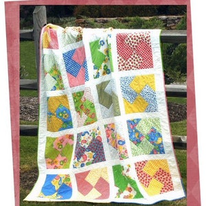 May include: A colorful quilt with a "Twisty Cakes" title. The quilt is made of squares with different floral and geometric patterns in shades of pink, blue, green, yellow, and white. The quilt is hanging on a wooden fence.
