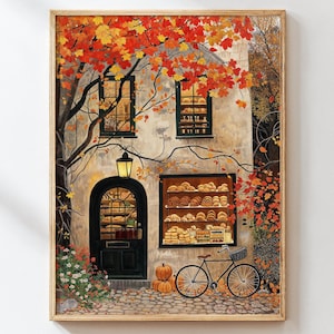 May include: Framed artwork depicting a bakery with a black door and a display window filled with baked goods. A bicycle with a basket is parked in front. The scene is set in autumn, with orange and red leaves.