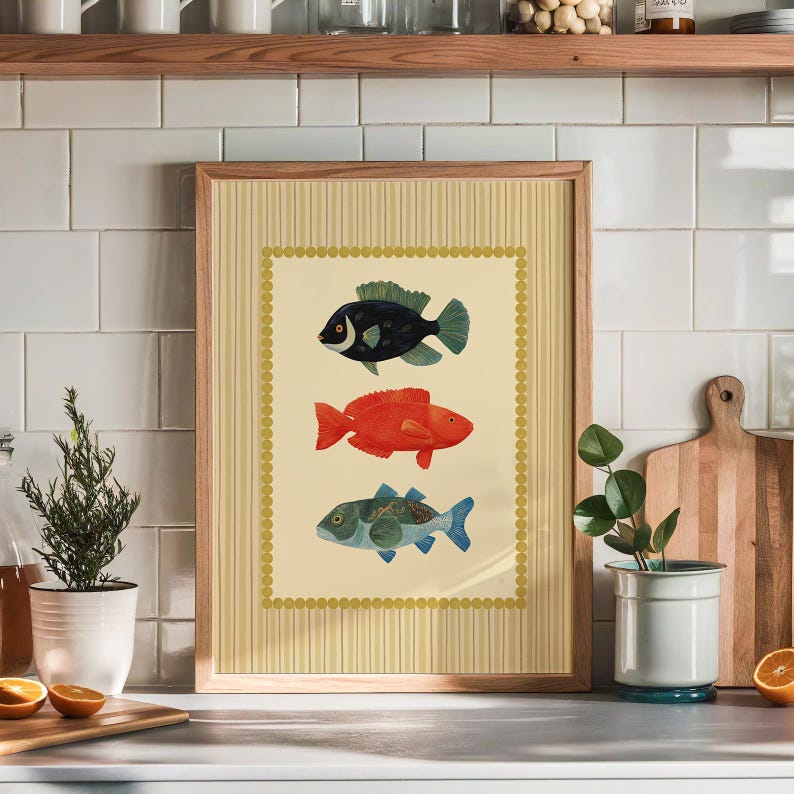 May include: A framed art print featuring three colorful fish illustrations. The fish are in black, red, and blue-green, set against a cream background with a gold border. The frame is light brown.