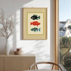 May include: A framed art print featuring three colorful fish illustrations. The fish are black, red, and blue-green, set against a cream background with a decorative border. The frame is a light wood color.