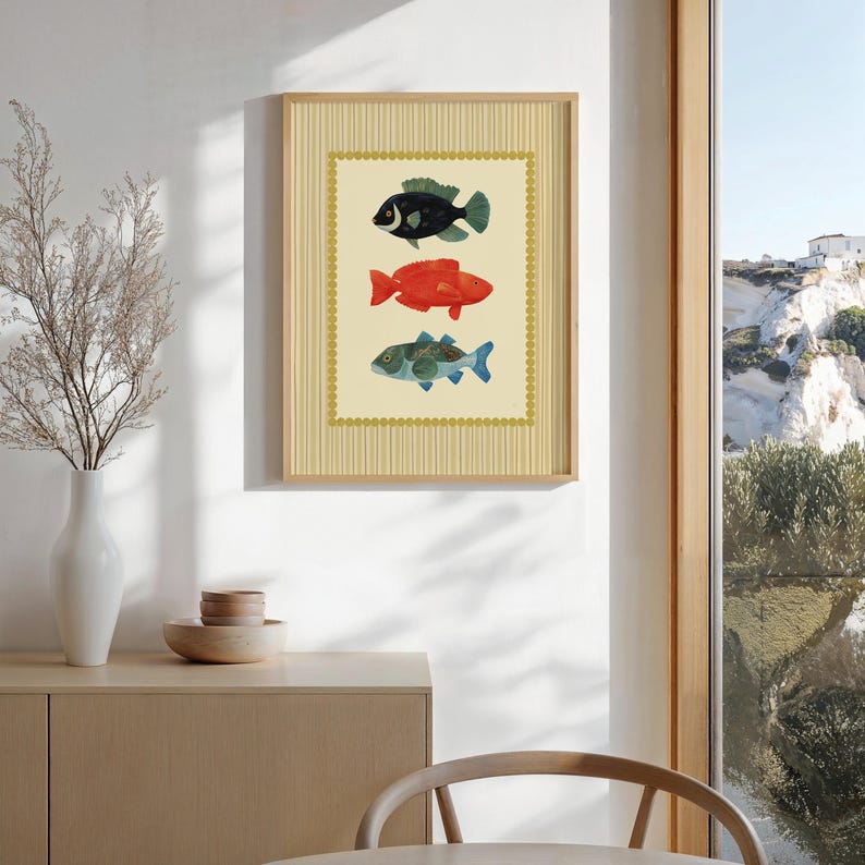 May include: A framed art print featuring three colorful fish illustrations. The fish are black, red, and blue-green, set against a cream background with a decorative border. The frame is a light wood color.