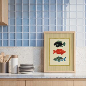 May include: A framed art print featuring three colorful fish illustrations. The fish are black, red, and blue-green, set against a cream background with a striped border. The frame is light wood, and the artwork is displayed on a countertop.
