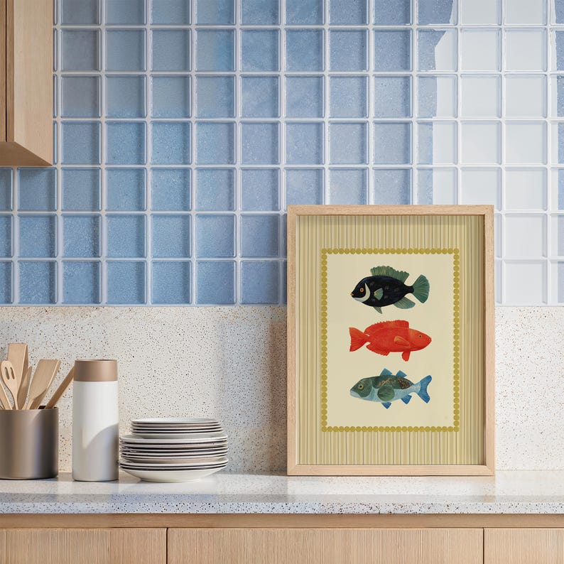 May include: A framed art print featuring three colorful fish illustrations. The fish are black, red, and blue-green, set against a cream background with a striped border. The frame is light wood, and the artwork is displayed on a countertop.