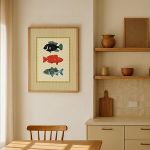 May include: Framed artwork featuring three colorful fish illustrations. The fish are in shades of black, red, and blue-green, set against a cream background with a vertical striped pattern. The frame is a light wood color.