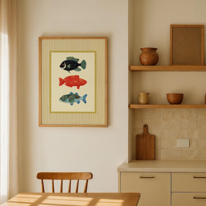 May include: Framed artwork featuring three colorful fish illustrations. The fish are in shades of black, red, and blue-green, set against a cream background with a vertical striped pattern. The frame is a light wood color.