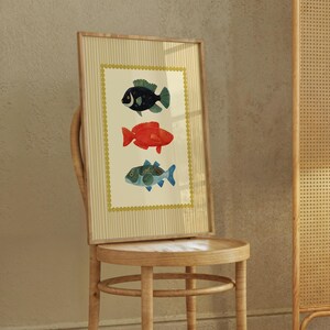 May include: A framed art print featuring three colorful fish illustrations. The fish are in shades of black, red, and blue-green, set against a cream background with a gold border. The frame is a light wood color.