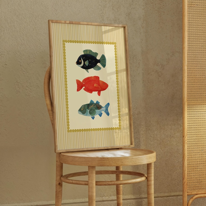 May include: A framed art print featuring three colorful fish illustrations. The fish are in shades of black, red, and blue-green, set against a cream background with a gold border. The frame is a light wood color.