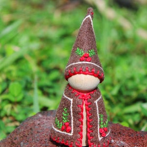 May include: A brown felt gnome with a red and white embroidered hat and a brown felt cape with red trim and green and red embroidered holly leaves.