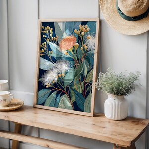 Puede incluir: Una impresi&oacute;n bot&aacute;nica enmarcada con un arreglo floral estilizado con flores naranjas, blancas y verdes sobre un fondo azul oscuro. La obra de arte se exhibe en un banco de madera, con un sombrero de paja y un jarr&oacute;n cerca.