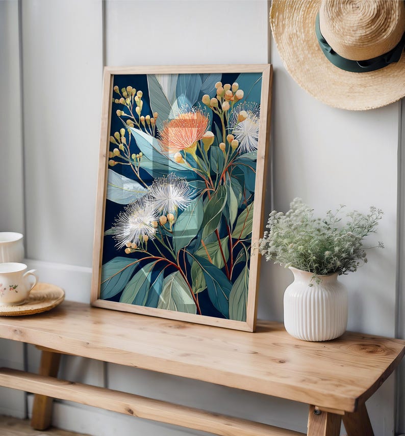 Puede incluir: Una impresi&oacute;n bot&aacute;nica enmarcada con un arreglo floral estilizado con flores naranjas, blancas y verdes sobre un fondo azul oscuro. La obra de arte se exhibe en un banco de madera, con un sombrero de paja y un jarr&oacute;n cerca.