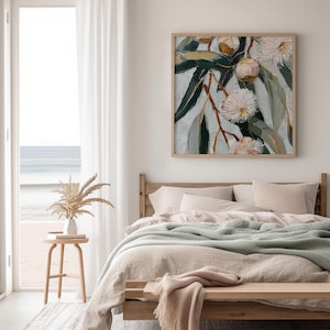 May include: A framed artwork depicting a close-up of eucalyptus branches with green leaves and white, fluffy flowers. The painting has a textured, impressionistic style. The artwork hangs above a bed with neutral-toned bedding and a light green blanket.