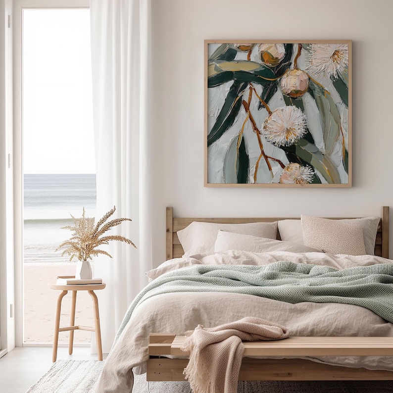 May include: A framed artwork depicting a close-up of eucalyptus branches with green leaves and white, fluffy flowers. The painting has a textured, impressionistic style. The artwork hangs above a bed with neutral-toned bedding and a light green blanket.
